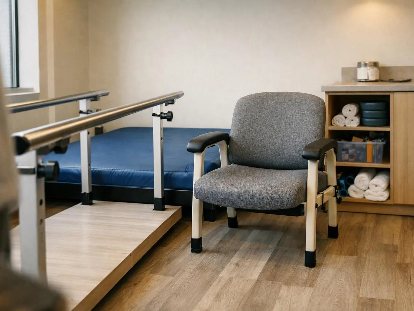 A gray chair sits beside a blue therapy bed, with parallel bars and folded towels in a calm room
