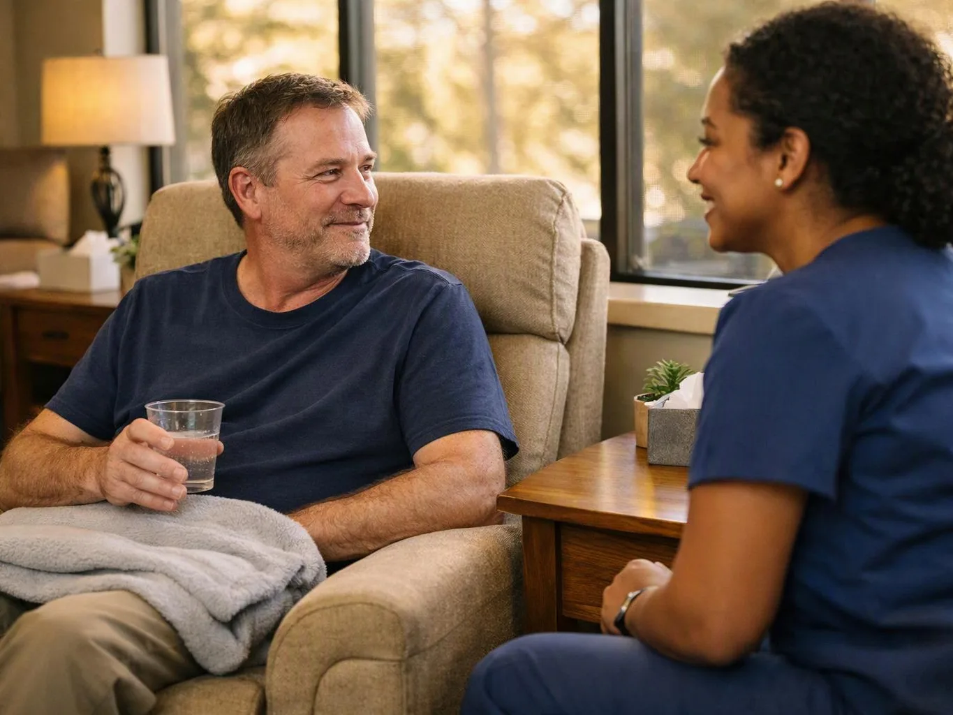 A man in an armchair holds a cup while smiling at a nurse in blue scrubs beside him