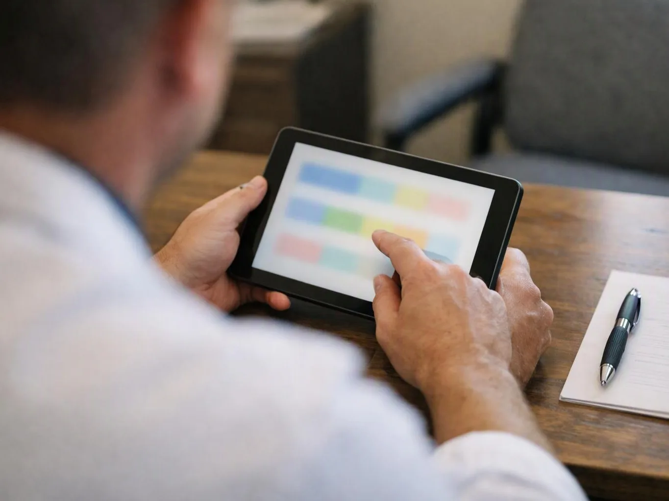 A man taps a tablet with blurred colorful blocks while sitting at a wooden desk with a pen and notepad