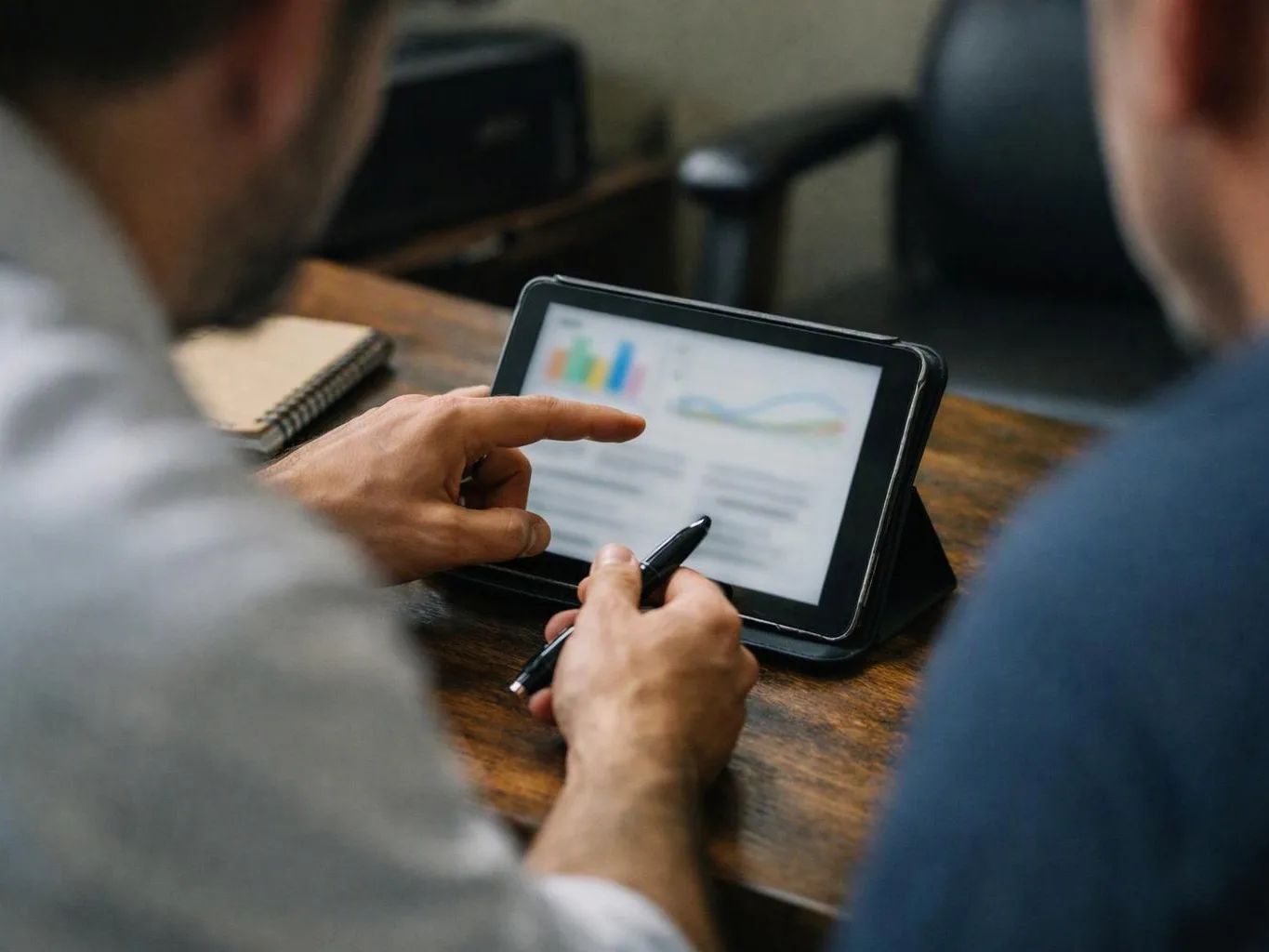 Two people study a tablet showing charts while one points and holds a pen at a wooden table